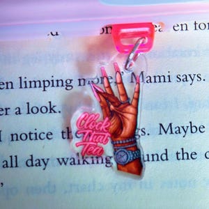 May include: A clear acrylic bookmark with a pink clip, featuring a hand illustration with long pink nails, a watch, and the text "Clock That Tea". The bookmark is attached to a book.