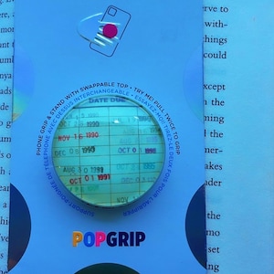 Peut inclure: Emballage bleu pour support de téléphone PopSockets avec le nom de la marque en lettres colorées. Une loupe montre une liste de dates. Le texte dit "Support-poignée de téléphone avec dessus interchangeable."