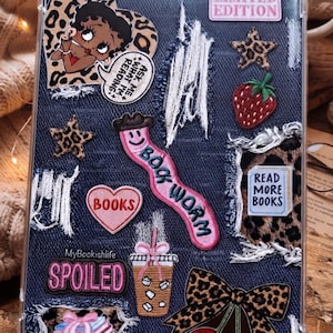 May include: A clear tablet case featuring a dark blue denim-textured background with distressed white frayed edges. It is decorated with various stickers including Betty Boop, leopard print stars, a strawberry, a pink bookworm patch, and text reading Limited Edition, Ask Me What I'm Reading, Books, Read More Books, MyBookishlife Spoiled, and a coffee cup.