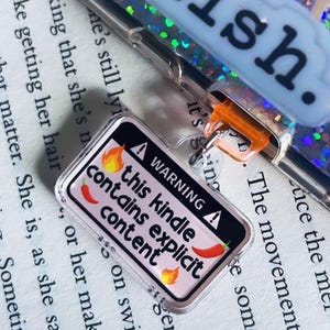 Könnte beinhalten: Ein rosa-schwarzes Warnschild mit dem Text "WARNING this kindle contains explicit content" und einer roten Chilischote. Das Schild ist an einem durchsichtigen Plastik-Schlüsselanhänger mit einem silbernen Ring befestigt.