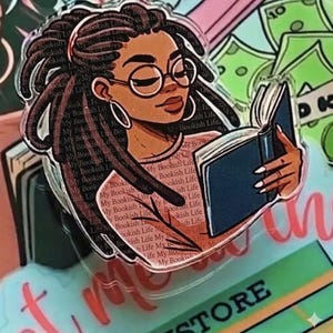 May include: A clear acrylic pin featuring a Black woman with locs, wearing glasses, and reading a book. The pin has the text "My Bookish Life" repeated throughout the design. The background includes images of money and books.