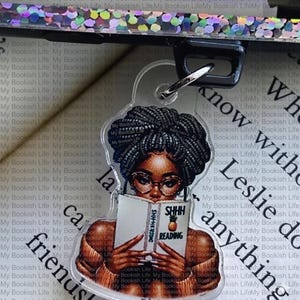 Pode incluir: Chaveiro de acrílico transparente com uma mulher negra de óculos lendo um livro com "SHHH READING" na capa. O chaveiro tem um fecho prateado e as palavras "My Bookish Life" são repetidas no fundo.