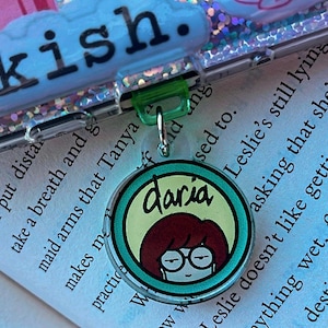 May include: A green and silver clip with a round, turquoise-colored charm featuring a cartoon image of Daria Morgendorffer from the animated television series Daria. The charm has a yellow background with the word "Daria" written in black cursive.