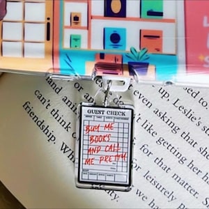 Puede incluir: Un llavero acrílico transparente con un cierre de metal plateado. El llavero presenta un diseño de "Guest Check" en miniatura con texto rojo escrito a mano que dice "BUY ME BOOKS AND CALL ME PRETTY". El fondo incluye un libro y una estantería colorida.