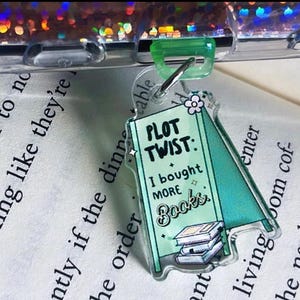 Pode incluir: Um chaveiro de acrílico verde e branco com uma placa que diz "Plot Twist: I bought more books". A placa está apoiada em uma pilha de livros.