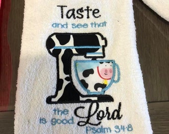 May include: White kitchen towel with an embroidered design of a black and white cow-print stand mixer and a blue and white cow-print cup. The text "Taste and see that the Lord is good Psalm 34:8" is embroidered in blue thread.