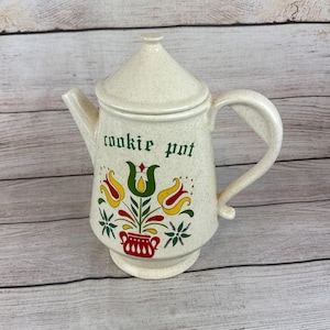 Puede incluir: Bote de galletas de cerámica color crema con tapa y asa. El bote presenta un diseño floral en amarillo, rojo y verde, con las palabras "cookie pot" en letras verdes. El bote tiene un acabado moteado y una base redondeada.