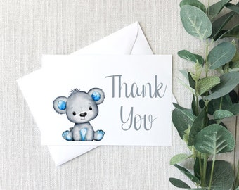 May include: A thank you card with a gray teddy bear with blue ears and a blue diamond on its chest. The text "Thank You" is written in gray on the card.
