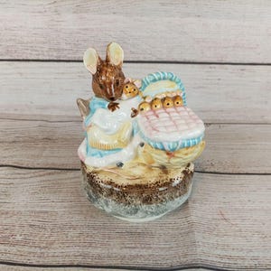 Puede incluir: Figura de cerámica de una madre ratón con sus bebés en un cochecito. El ratón es marrón y lleva un vestido azul y blanco. El cochecito es rosa y azul, y la base es marrón y beige.