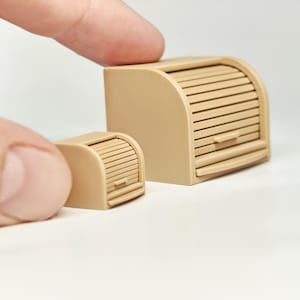 May include: Two miniature tan-colored bread boxes with a roll-top design. The larger box has a handle and horizontal slats. The smaller box mirrors the design. The boxes are on a white surface.