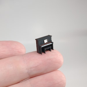 Op de afbeelding: Miniatuur zwart piano met een bijpassende bank. De kleine piano heeft witte toetsen en een bladmuziek op de standaard. Een kleine, gedetailleerde replica van een vleugel, ideaal voor poppenhuizen of knutselen.