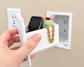 May include: A white rectangular wall safe designed to look like an electrical outlet cover. The open container holds a black smartwatch, a gold chain, and a small rolled item. The unit is being held in front of a matching wall-mounted frame.