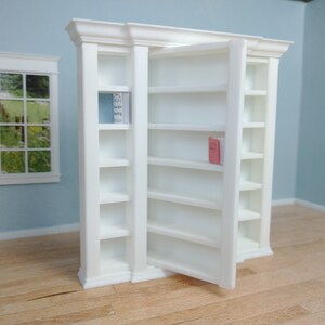May include: White bookcase with a door that swings open to reveal shelves. The bookcase has a decorative trim around the top and sides. The shelves are empty except for a single book on the top shelf.