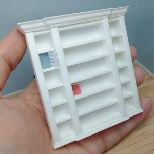May include: White miniature bookshelf with six shelves on each side. The shelves are empty except for a blue and white book on the top shelf of the left side and a pink book on the middle shelf of the left side.