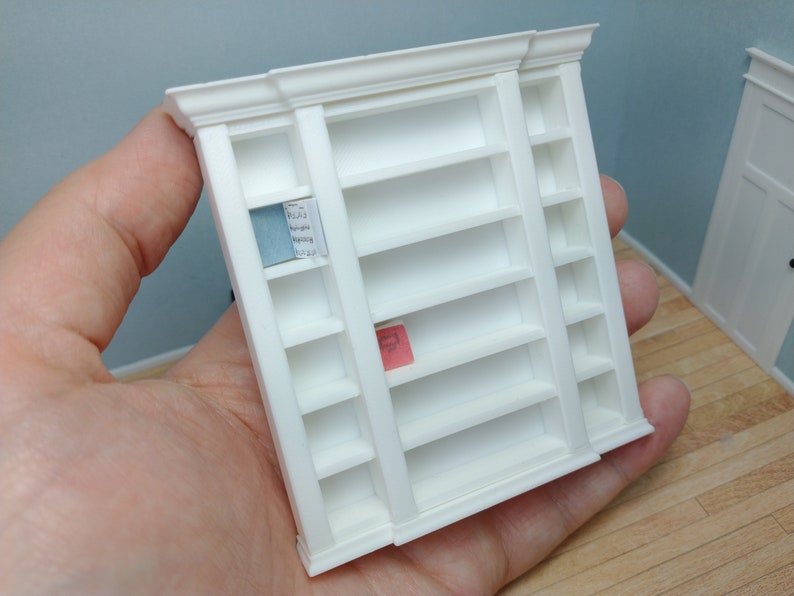 May include: White miniature bookshelf with six shelves on each side. The shelves are empty except for a blue and white book on the top shelf of the left side and a pink book on the middle shelf of the left side.