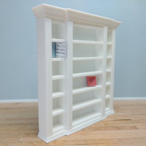 May include: White wooden bookcase with six shelves on each side and a single shelf in the middle. The bookcase has a decorative molding around the top and bottom. A small red book is on one of the shelves.