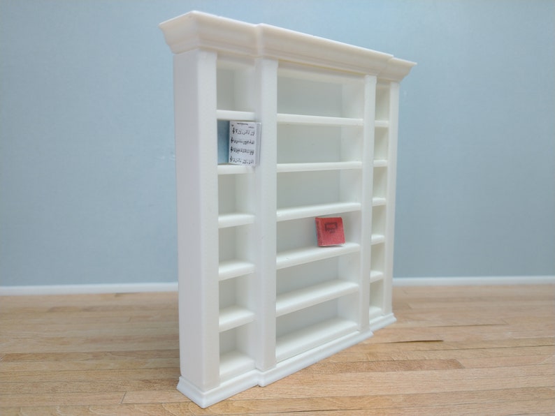 May include: White wooden bookcase with six shelves on each side and a single shelf in the middle. The bookcase has a decorative molding around the top and bottom. A small red book is on one of the shelves.