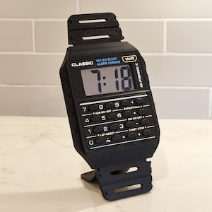 May include: A black digital wristwatch featuring a rectangular screen displaying the time 7:18 and a built-in calculator keypad below the display. The watch face includes the text CLASSIC, WATER RESIST, ALARM CHRONO, and WR, with a black strap.