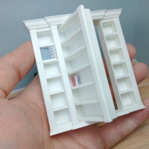 May include: White miniature bookshelf with a door that opens to reveal additional shelves. The bookshelf has a decorative trim around the top.