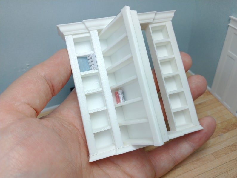 May include: White miniature bookshelf with a door that opens to reveal additional shelves. The bookshelf has a decorative trim around the top.