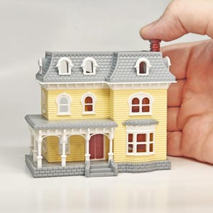 May include: A miniature yellow Victorian house with a gray roof, porch, and trim. The house has arched windows, a red door, and a brick chimney. The house is held by a hand.