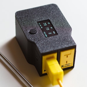 Puede incluir: Term&oacute;metro digital negro y amarillo con una sonda de acero inoxidable. El dispositivo muestra lecturas en una peque&ntilde;a pantalla, mostrando "73 73 2" y "11:16 PM". El texto "CHROM + ALUMEL" es visible.