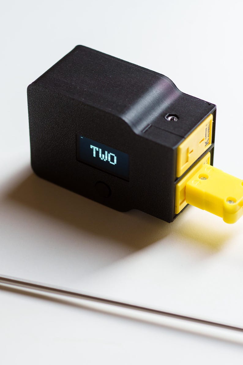 Puede incluir: Dispositivo electr&oacute;nico negro con conector amarillo y la palabra &laquo;TWO&raquo; en una peque&ntilde;a pantalla. El dispositivo tiene una superficie texturizada, probablemente para medici&oacute;n o monitoreo.