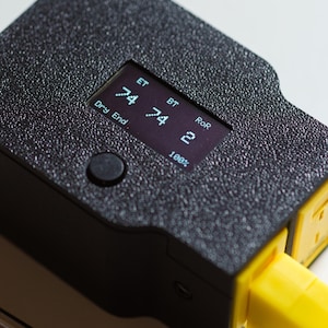 Puede incluir: Primer plano de un dispositivo electr&oacute;nico negro con una superficie texturizada. El dispositivo tiene una peque&ntilde;a pantalla que muestra n&uacute;meros y texto, incluyendo "ET", "BT" y "RoR". Un enchufe amarillo se extiende desde el lateral. Un bot&oacute;n es visible.