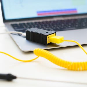 Peut inclure: Un appareil rectangulaire noir avec un connecteur jaune est branché sur un ordinateur portable. Un câble jaune enroulé part de l'appareil. L'ordinateur portable est ouvert sur une surface blanche.