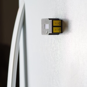 Puede incluir: Un peque&ntilde;o dispositivo rectangular con una carcasa de metal gris plateado y dos paneles amarillos. El dispositivo est&aacute; adherido a una superficie blanca, posiblemente la puerta de un refrigerador. Los paneles amarillos tienen texto y s&iacute;mbolos negros.