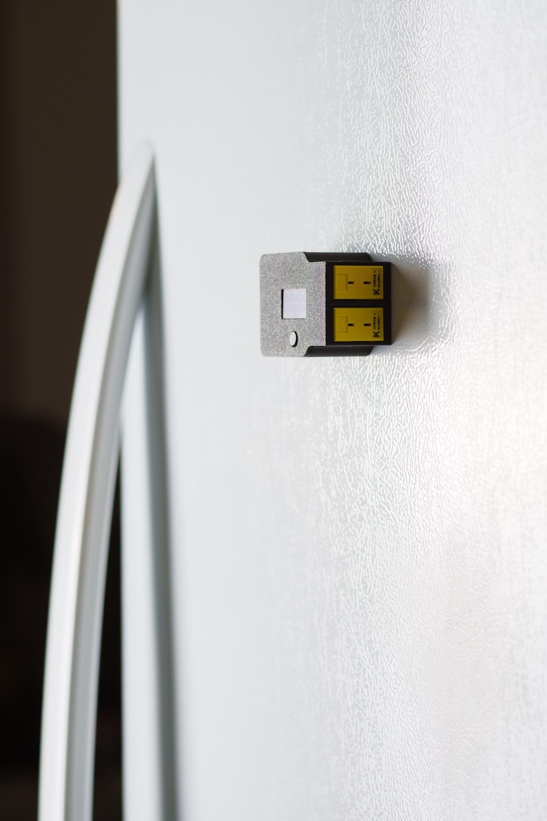 Puede incluir: Un peque&ntilde;o dispositivo rectangular con una carcasa de metal gris plateado y dos paneles amarillos. El dispositivo est&aacute; adherido a una superficie blanca, posiblemente la puerta de un refrigerador. Los paneles amarillos tienen texto y s&iacute;mbolos negros.