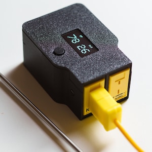 Puede incluir: Un term&oacute;metro digital negro con un enchufe amarillo y una sonda de acero inoxidable. La pantalla muestra la temperatura en formato digital. El term&oacute;metro est&aacute; dise&ntilde;ado para medir temperaturas.