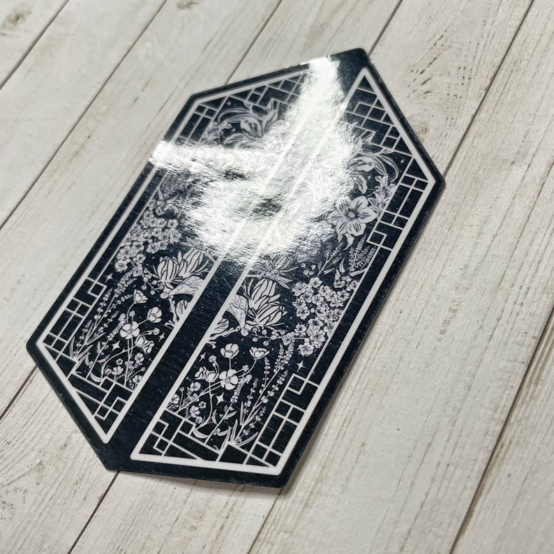 May include: Black and white sticker with a floral design inside a geometric frame. The sticker is shaped like a hexagon.