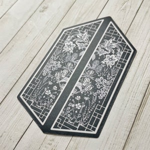 May include: Black and white sticker with a geometric design featuring a floral pattern. The sticker is shaped like a hexagon and has a black background with white flowers and leaves.