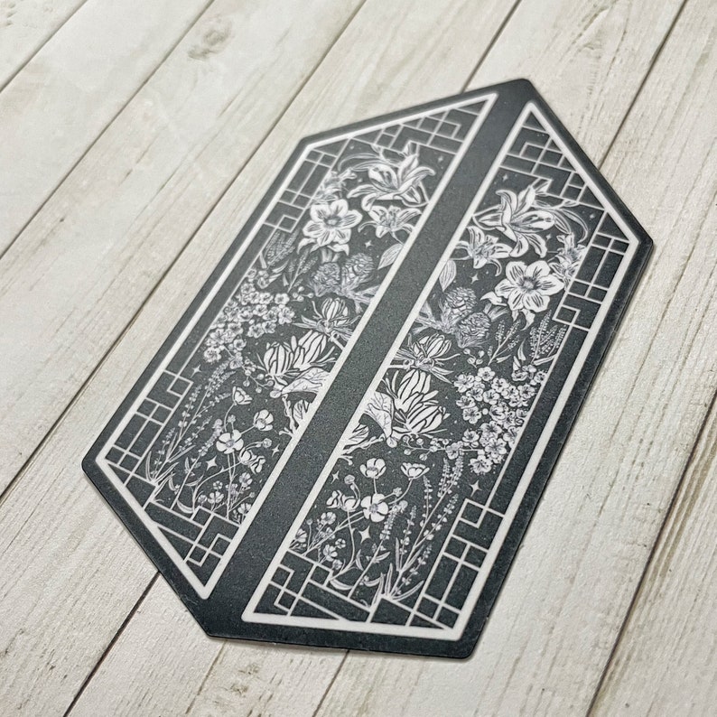May include: Black and white sticker with a geometric design featuring a floral pattern. The sticker is shaped like a hexagon and has a black background with white flowers and leaves.