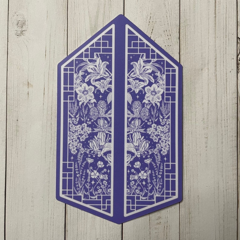 May include: A purple geometric card with a white floral design. The card features a symmetrical design with a floral pattern on each side. The flowers are stylised and include lilies, leaves, and other botanical elements.