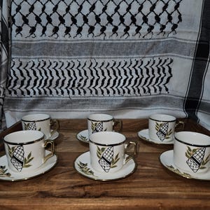 May include: A set of six white teacups and saucers with gold trim, arranged on a wooden tray. Each cup and saucer features a black and green geometric design. The background includes a black and white patterned textile.