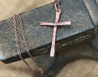 May include: A handcrafted copper cross pendant with a hammered texture, suspended from a delicate chain. The cross is displayed on a dark, textured surface, showcasing its rustic charm. The chain and cross have a warm, reddish-brown hue.