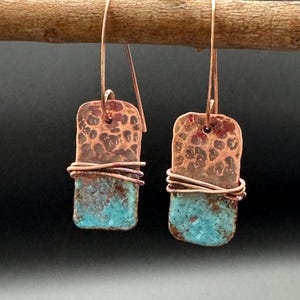 May include: A pair of handcrafted copper earrings. Each earring features a hammered copper top section and a turquoise-colored bottom section, wrapped with copper wire. The earrings hang from copper ear wires.