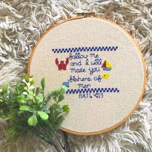 May include: A round, cross-stitched embroidery hoop with the text "Follow me and I will make you fishers of men" and the reference "MATT. 4:19". The design includes a crab, sailboat, and fish. The hoop is set against a textured background.