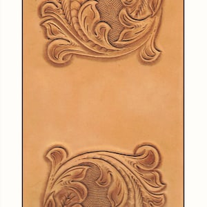 May include: Close-up of a piece of tan leather with two ornate, symmetrical floral designs tooled into the surface. The designs feature swirling leaves and textured backgrounds, creating a detailed, embossed effect.