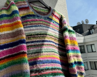 May include: A colourful striped knitted jumper with a round neckline. The jumper is hanging on a black hanger against a white wall.