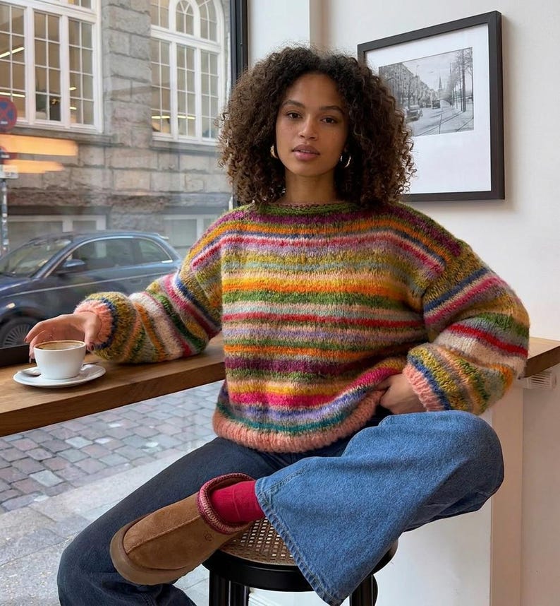 May include: A colorful, striped sweater with a round neckline. The sweater features horizontal bands of various colors, including green, orange, pink, and purple. The person is wearing blue jeans and brown boots. A cup of coffee sits on a wooden surface.