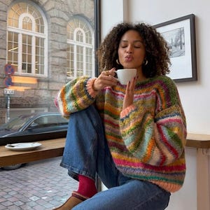 May include: A colorful, striped sweater with a round neckline. The sweater features a mix of pink, green, yellow, and blue horizontal stripes. The person is wearing blue jeans and red socks. A white coffee cup is held up to the person's lips.