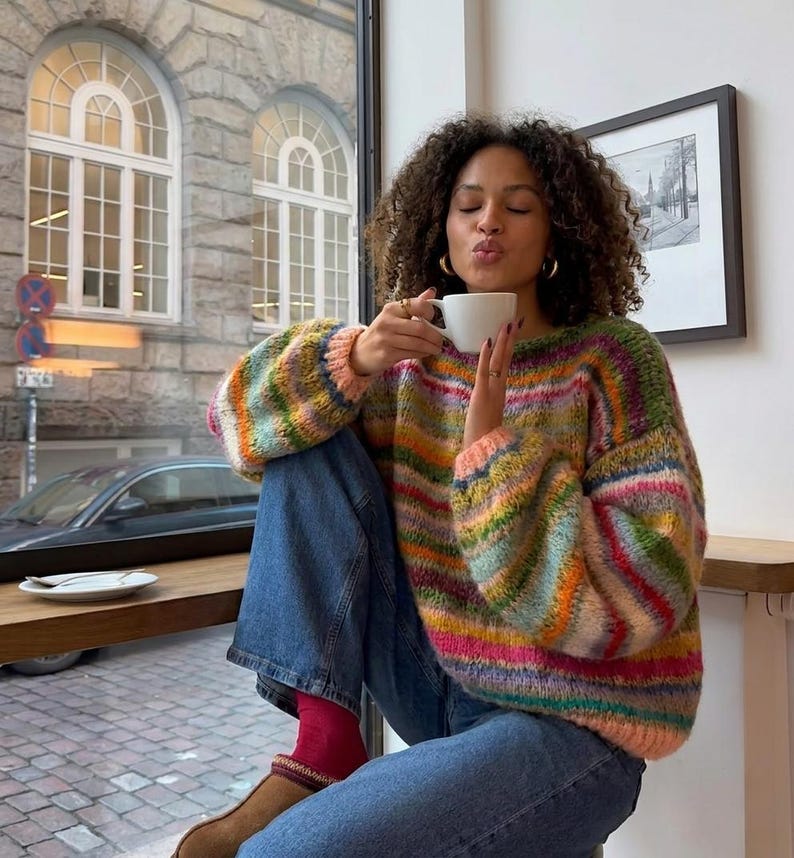 May include: A colorful, striped sweater with a round neckline. The sweater features a mix of pink, green, yellow, and blue horizontal stripes. The person is wearing blue jeans and red socks. A white coffee cup is held up to the person's lips.
