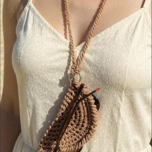 Puede incluir: Un estuche para gafas de macram&eacute; color beige con un collar de cord&oacute;n a juego. El estuche para gafas est&aacute; sujeto a un anillo plateado y contiene un par de gafas con montura marr&oacute;n. El collar se lleva sobre un vestido color crema.