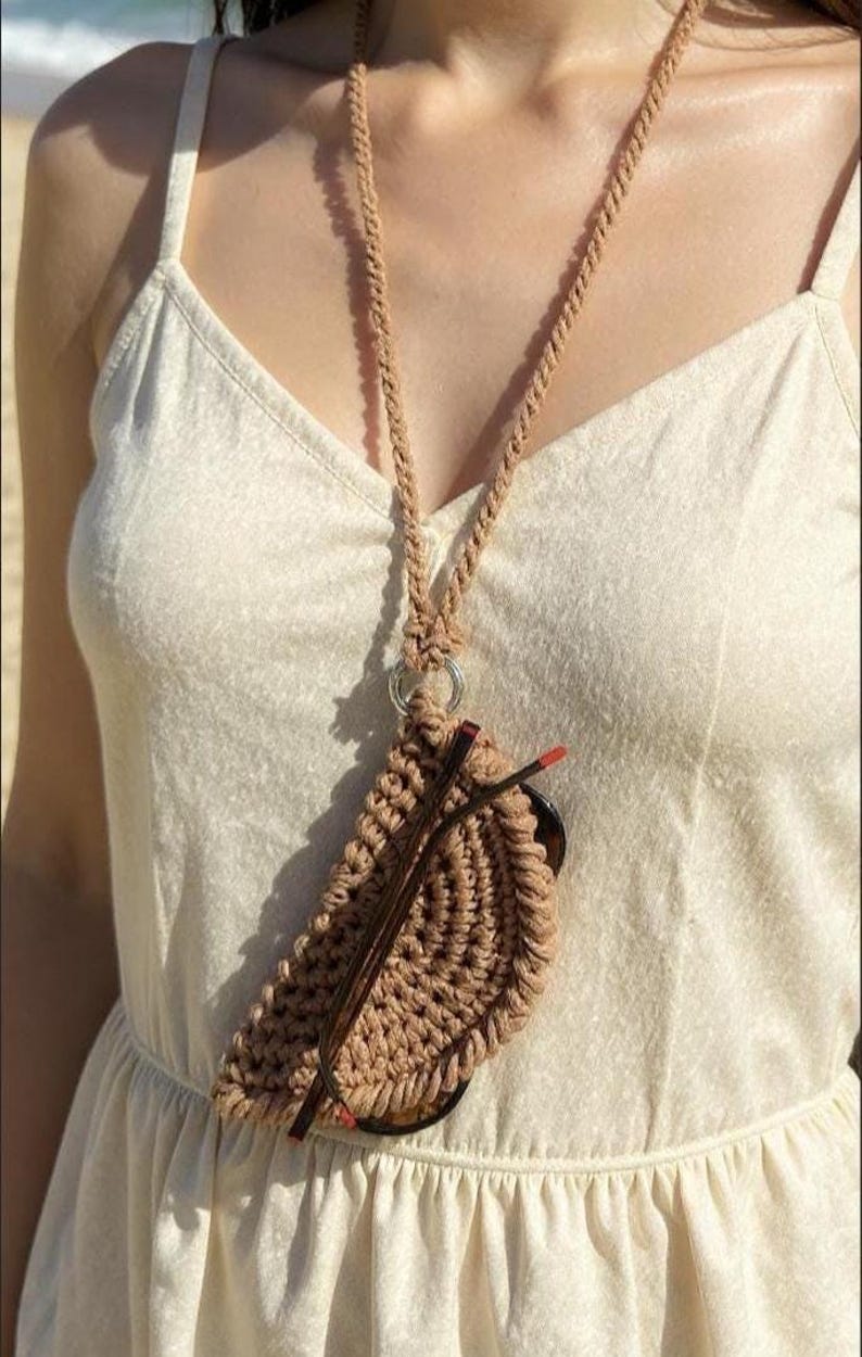 Puede incluir: Un estuche para gafas de macram&eacute; color beige con un collar de cord&oacute;n a juego. El estuche para gafas est&aacute; sujeto a un anillo plateado y contiene un par de gafas con montura marr&oacute;n. El collar se lleva sobre un vestido color crema.