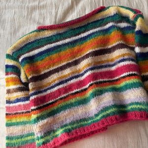 May include: A colorful, striped knit sweater with a round neckline and long sleeves. The sweater features a vibrant array of horizontal stripes in various colors, including pink, green, yellow, and brown. The neckline and cuffs are trimmed with a bright pink yarn.