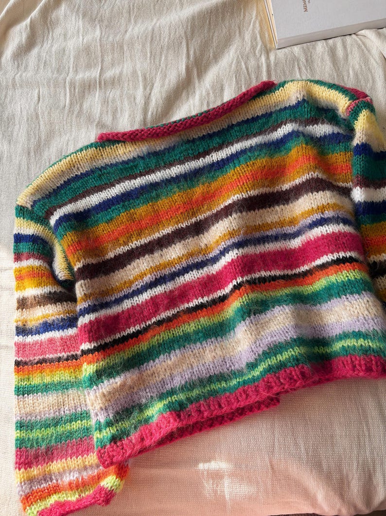 May include: A colorful, striped knit sweater with a round neckline and long sleeves. The sweater features a vibrant array of horizontal stripes in various colors, including pink, green, yellow, and brown. The neckline and cuffs are trimmed with a bright pink yarn.