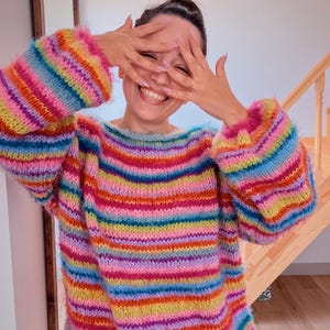 May include: A vibrant, striped sweater in pink, orange, yellow, green, blue, and purple. The sweater has long sleeves and a fluffy texture. The person in the image is smiling and covering their eyes with their hands.